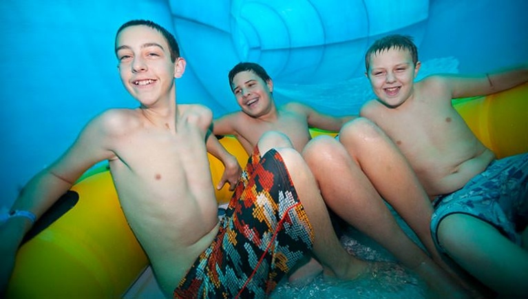 Three boys smile as they share a tube down the Double Whirlwind water slide at a Great Wolf Lodge indoor water park.