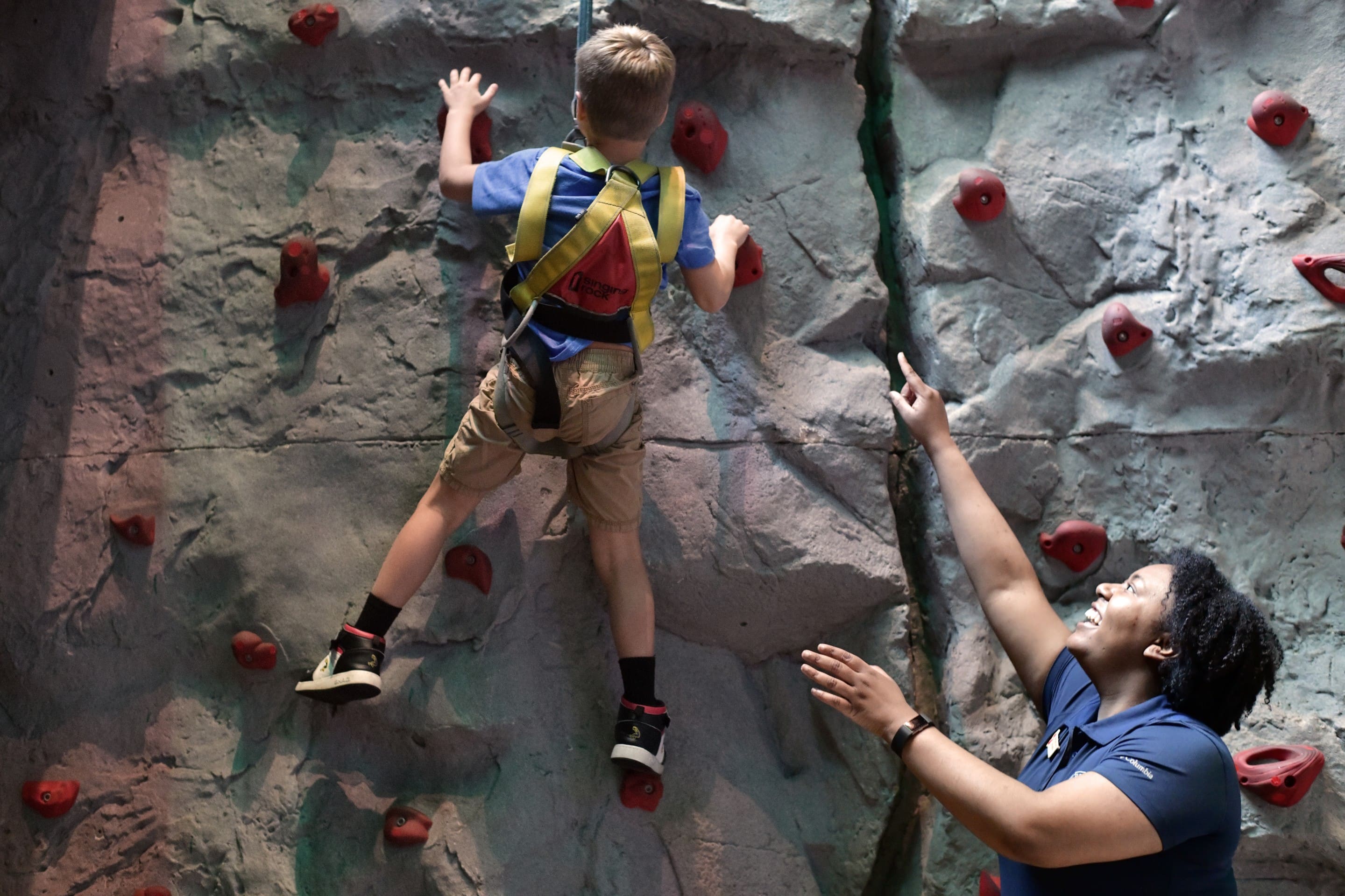 Child climbing rock wall while smiling instructor points upward for guidance