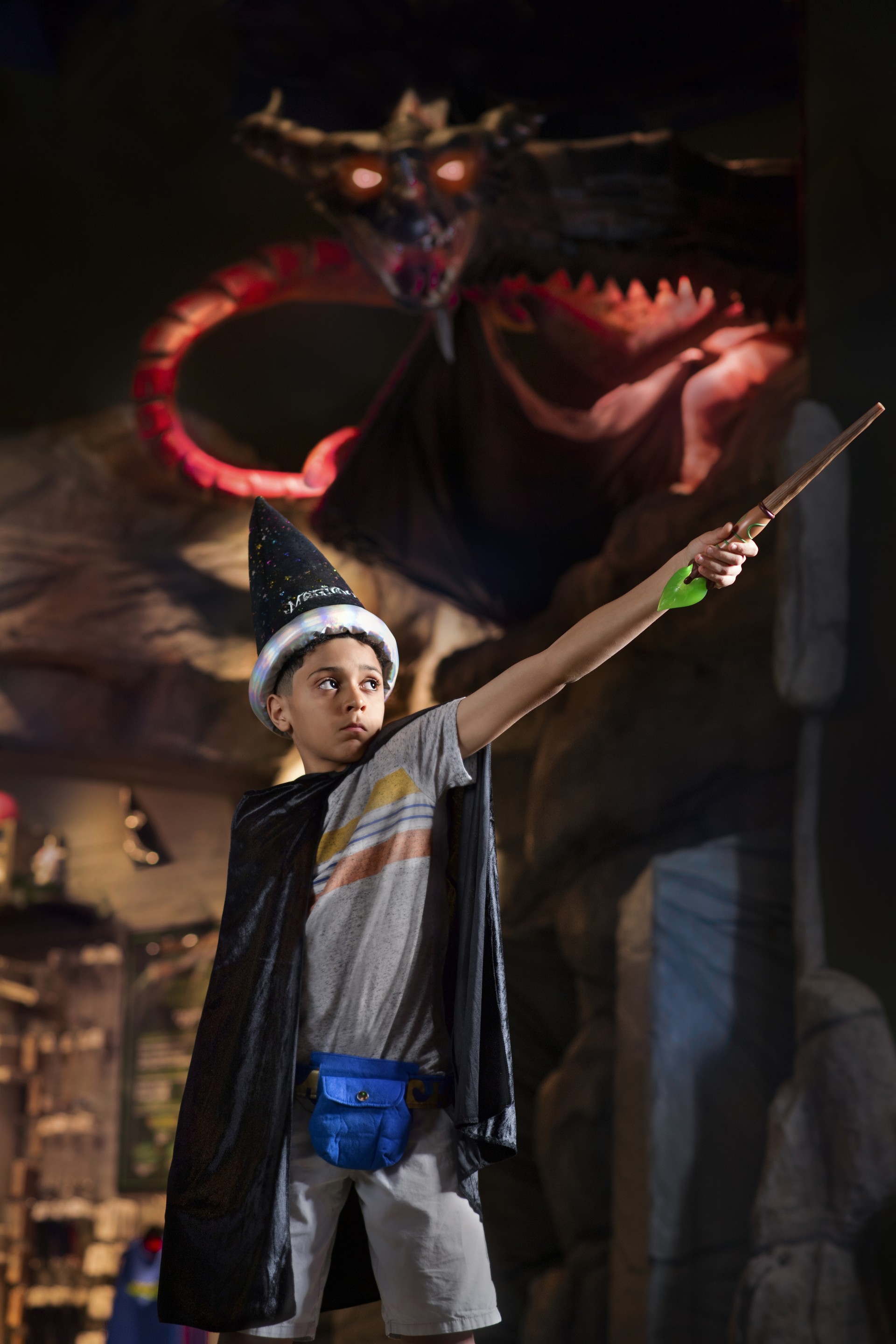 A young boy wearing a cape and sorcerer's hat with an outstretched arm holding a magic wand.
