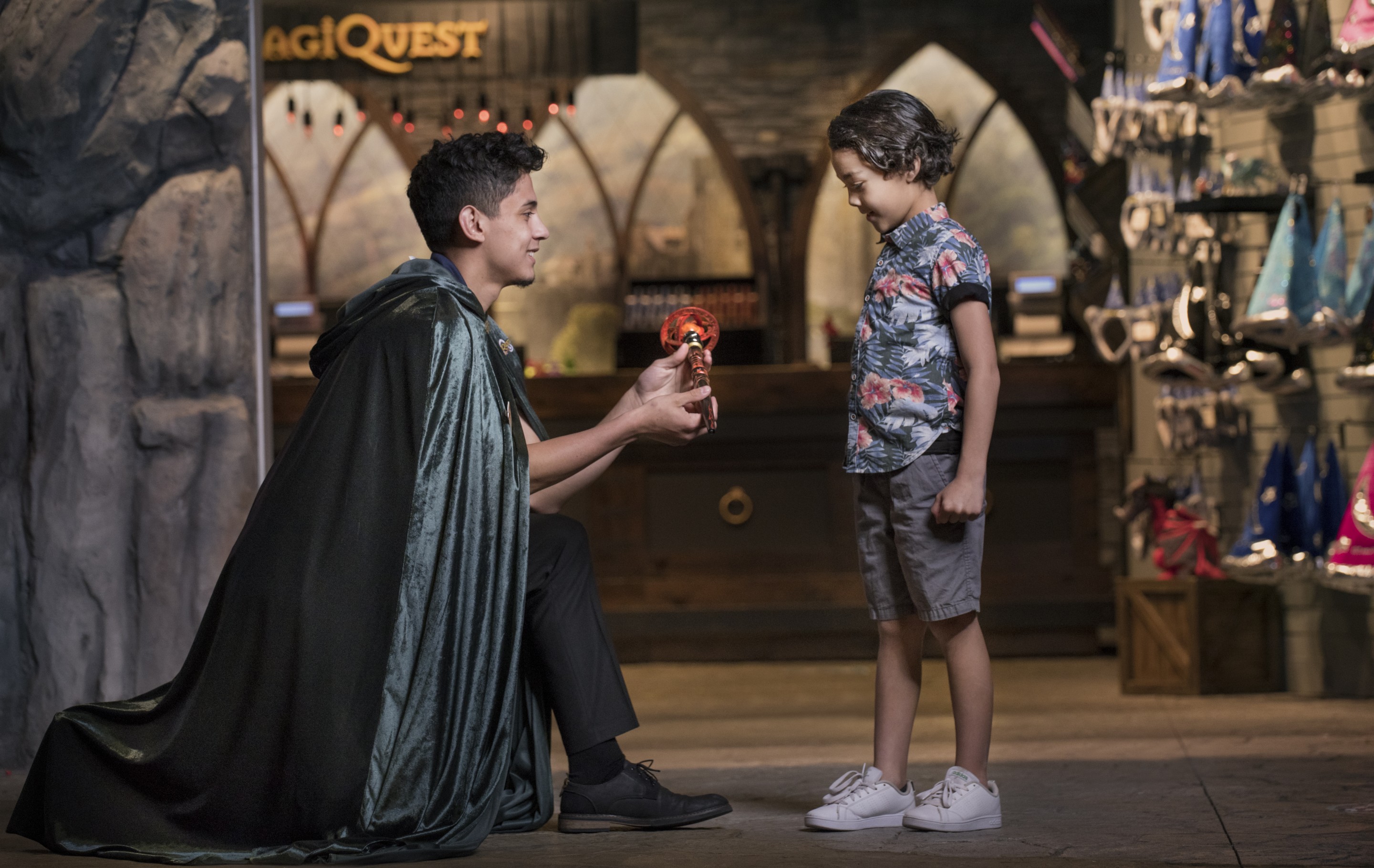 A teen boy wearing a cape kneeling down presenting a young boy with a MagiQuest toy.