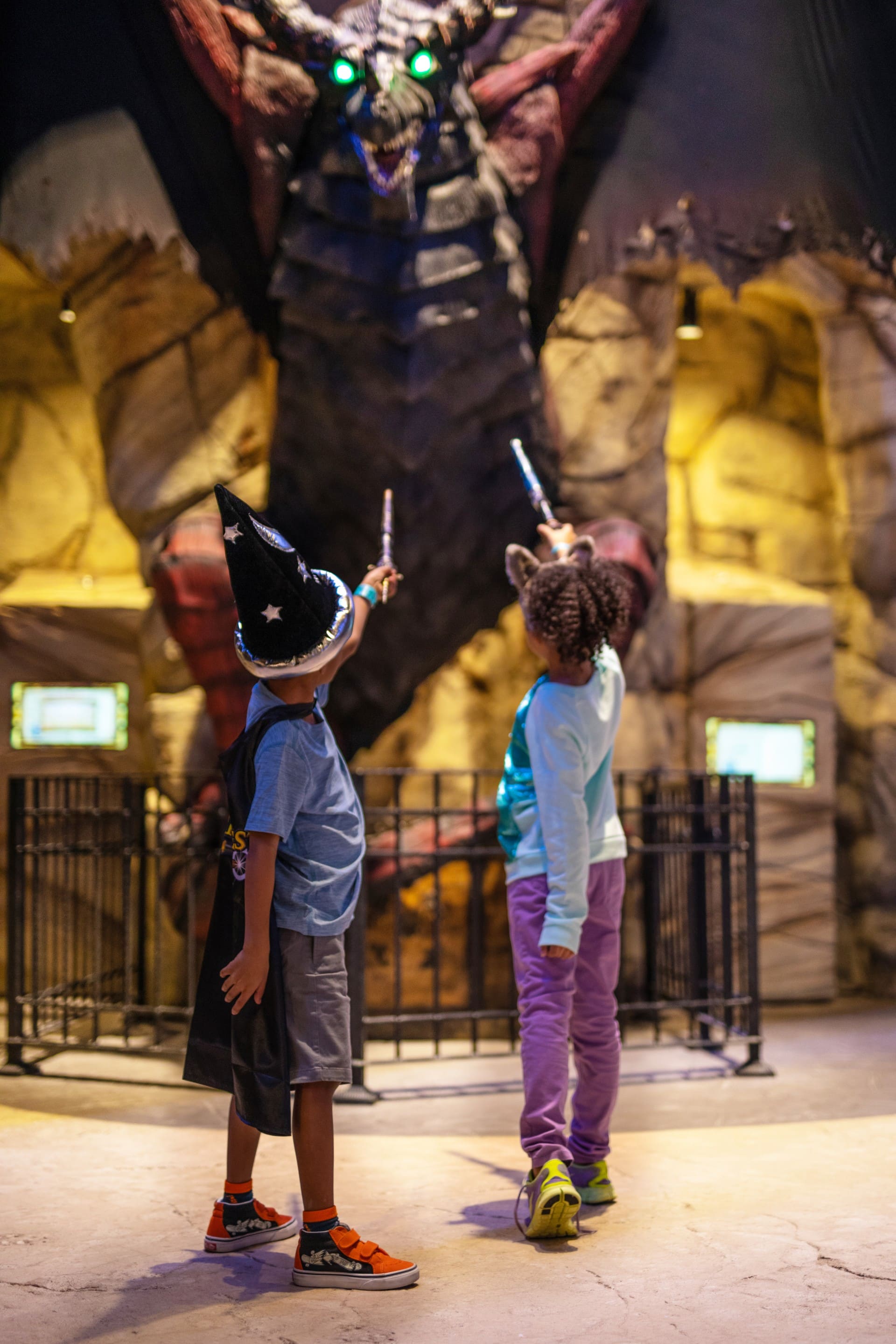 A young boy wearing a sorcerer's hat and a young girl point their wands at a MagiQuest Dragon..