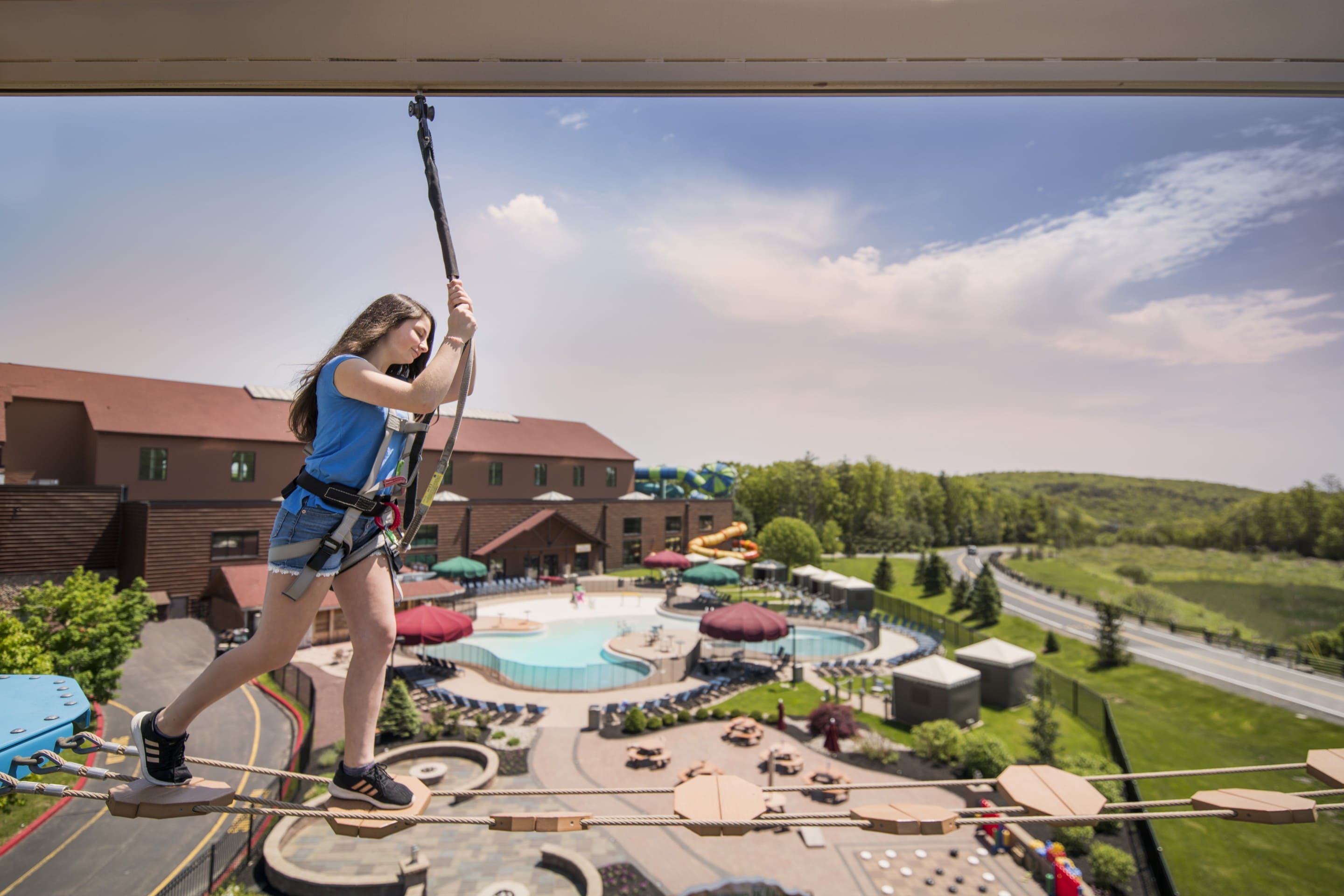 Girl in harness crossing ropes above outdoor pool area