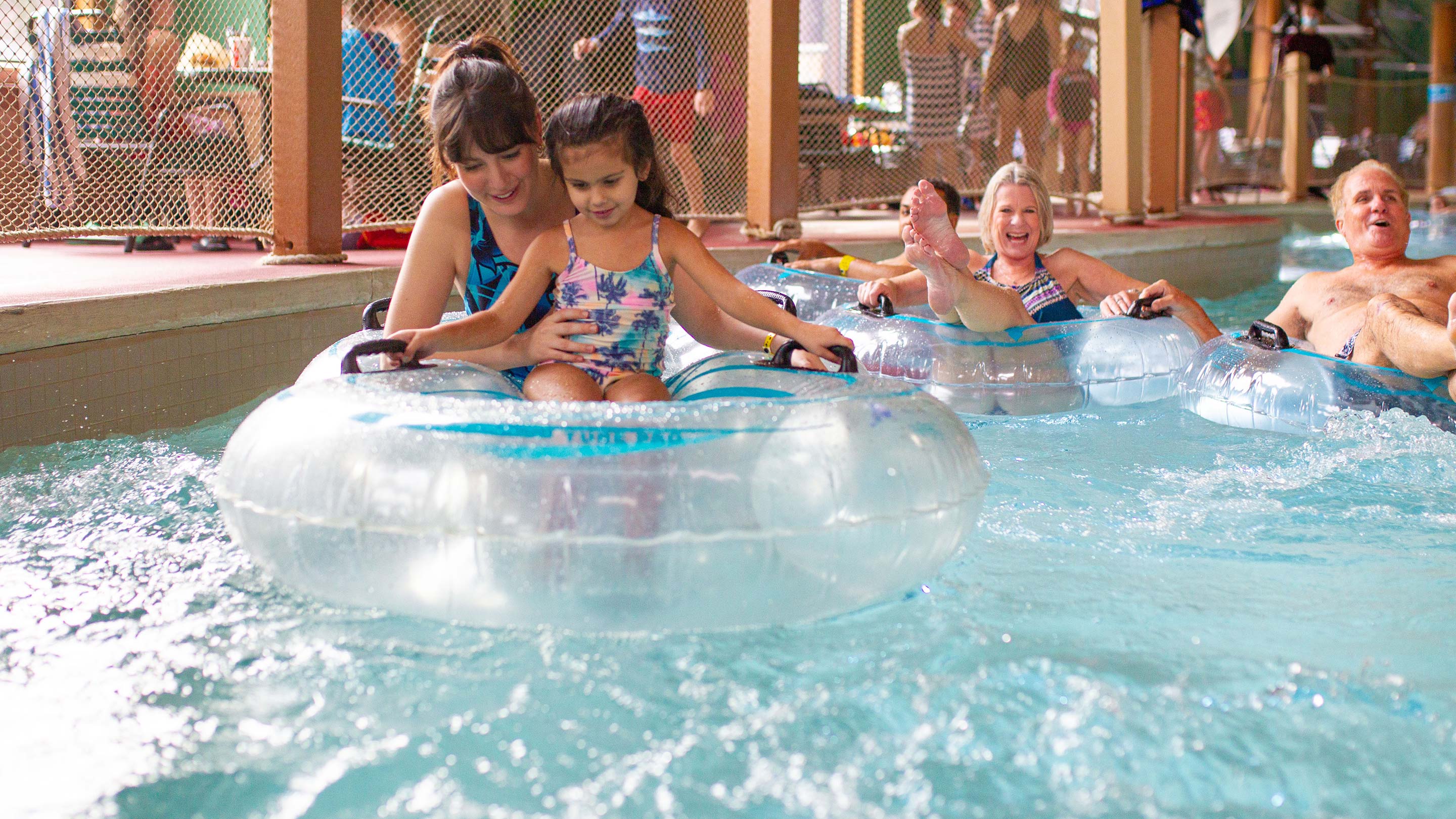 A mom in a clear inner-tube holding a child and enjoying the lazy river with group of adults in the background