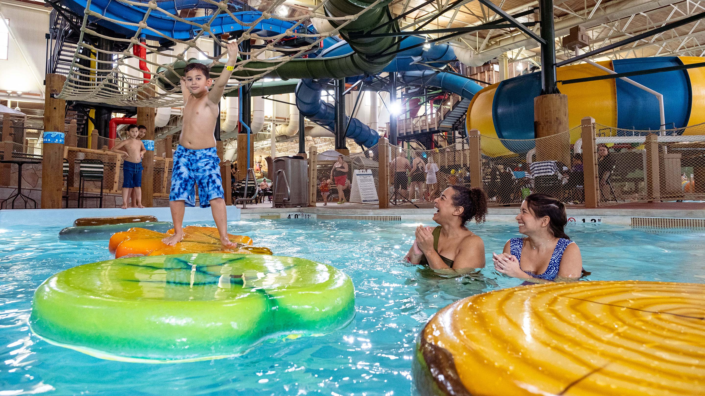 A young boy smiles as he grabs on to the rope net over a pool while balancing on inflatable lily rafts as two adult woman look on cheering.