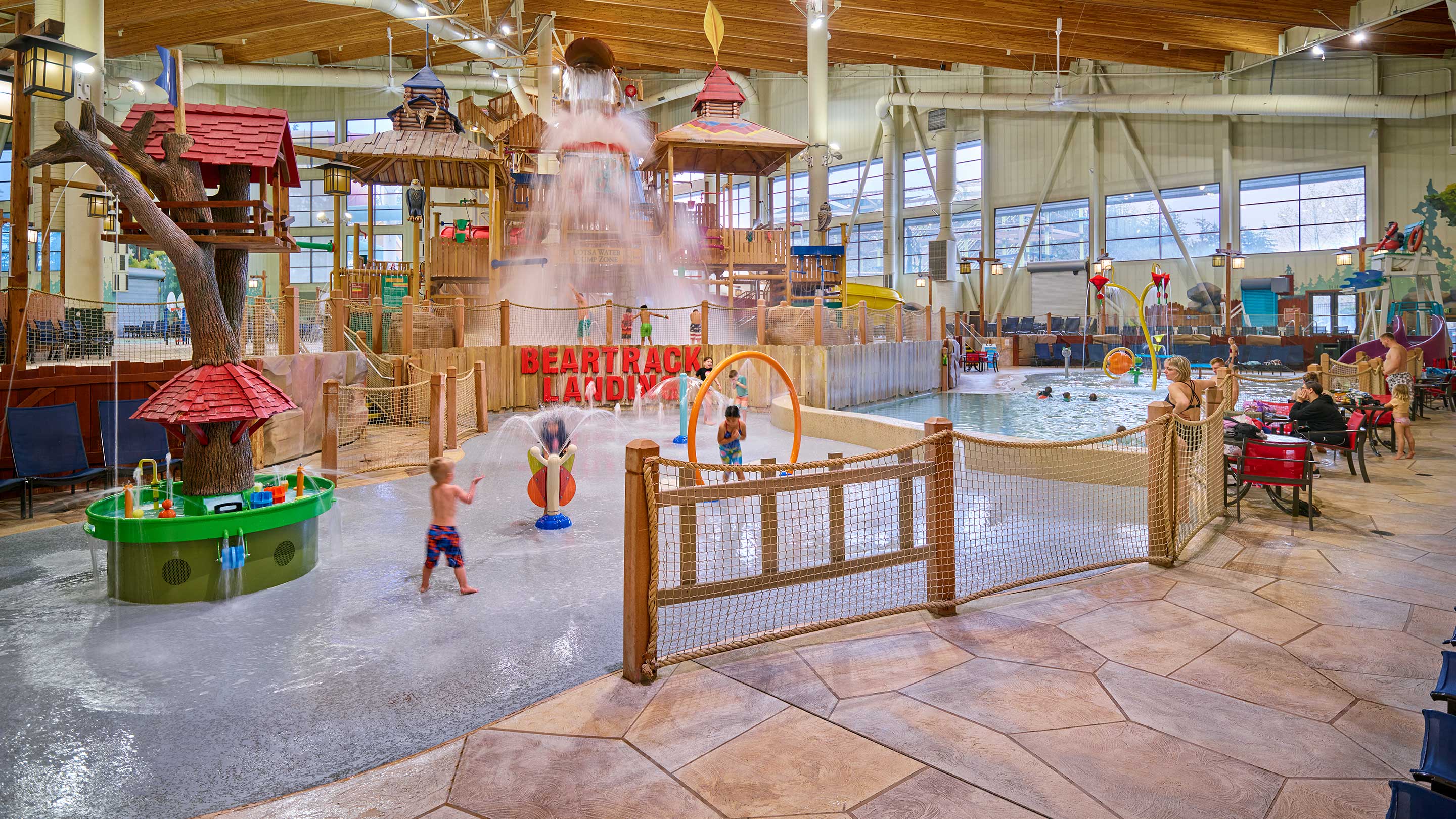 Kids are joyfully playing under a water feature at an indoor water park, surrounded by colorful play structures.